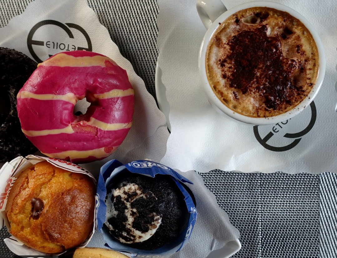 Cappuccino spolverato di cacao, ciambella glassata rosa e due muffin, uno Oreo, su tovaglioli col logo Etoile