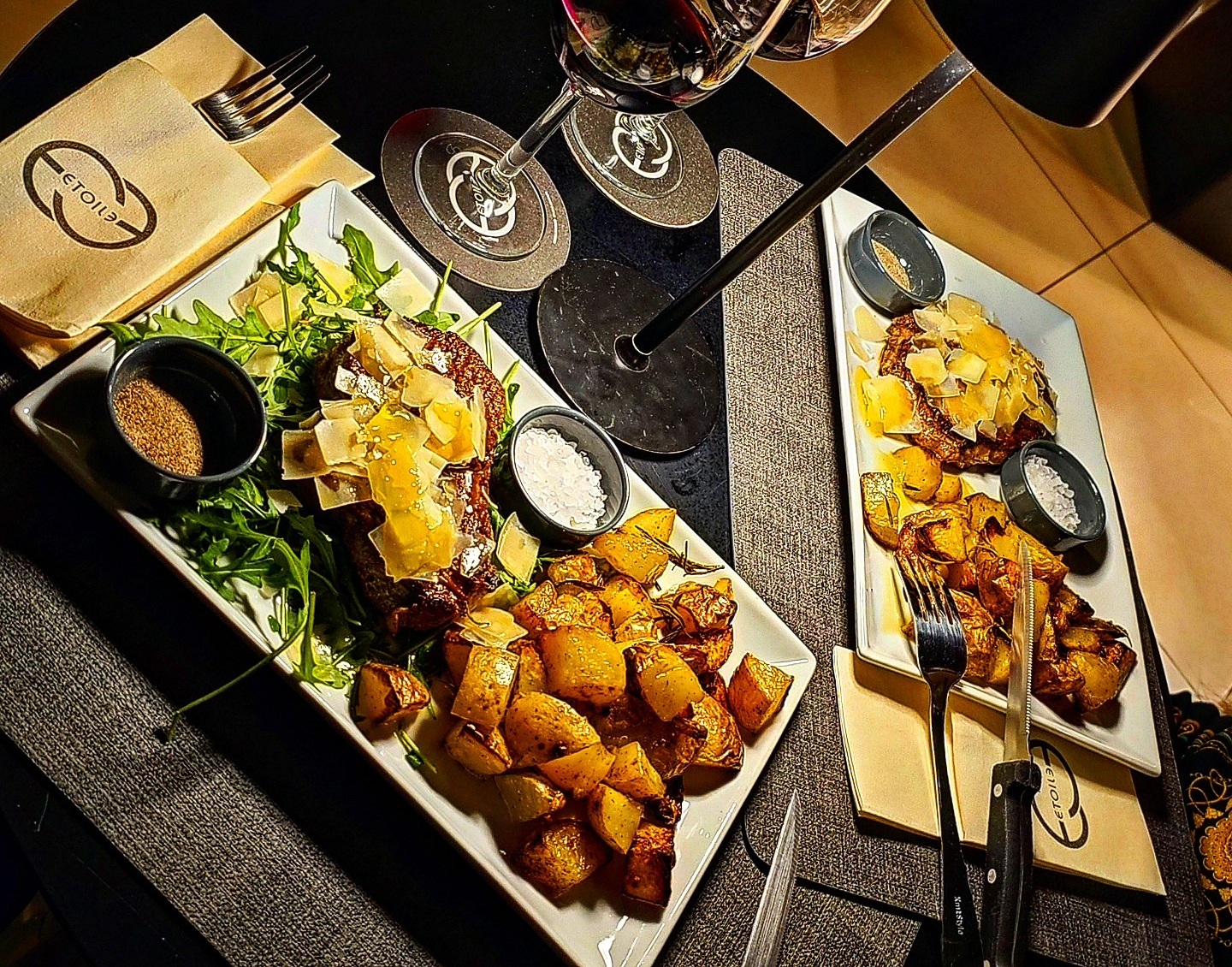 Due piatti di tagliata con scaglie di grana, rucola e patate al forno su tavolo apparecchiato con tovaglioli Etoile
