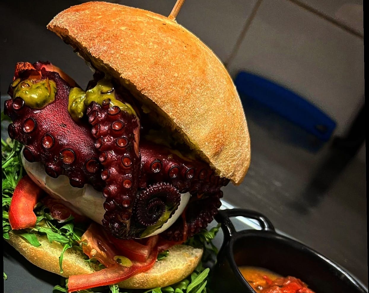 Panino con tentacoli di polpo scottati, mozzarella, pesto verde, pomodoro e rucola, accanto a un pentolino di sugo rosso