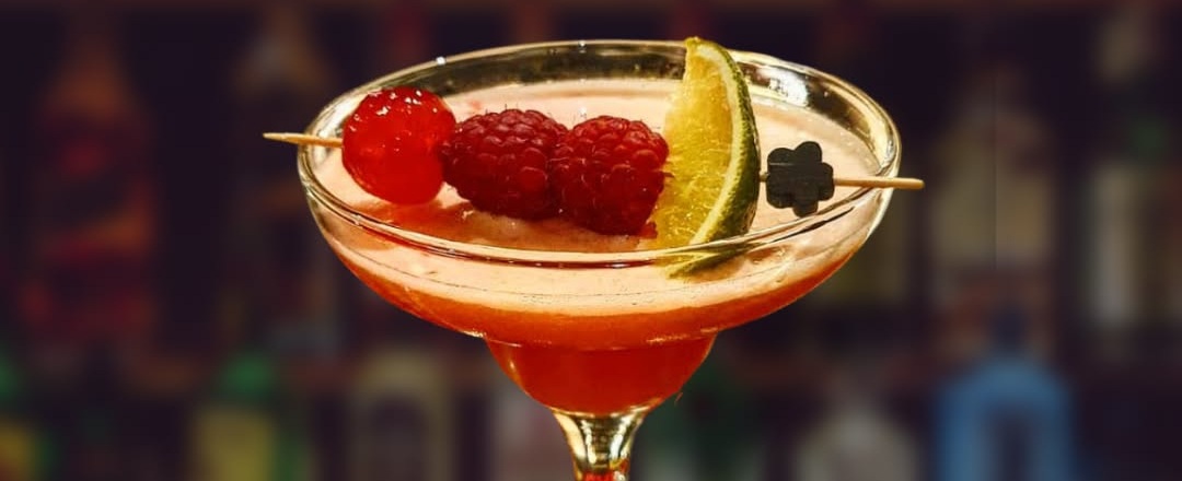 Cocktail rosso in coppa con schiuma, spiedino di lamponi, ciliegia e spicchio di lime, bottiglie sfocate sullo sfondo