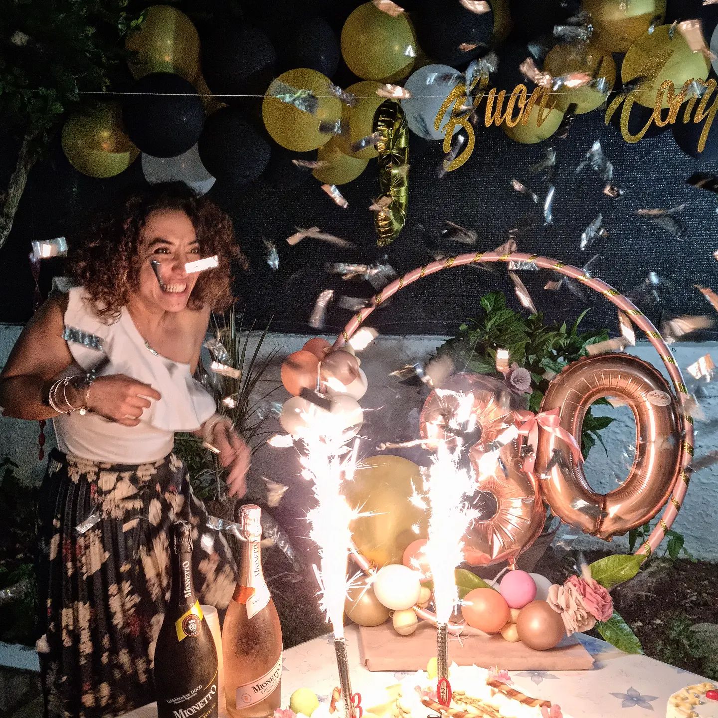 Donna sorridente tra coriandoli davanti a torta con fontane scintillanti, palloncini neri e oro e numero 30 rosa